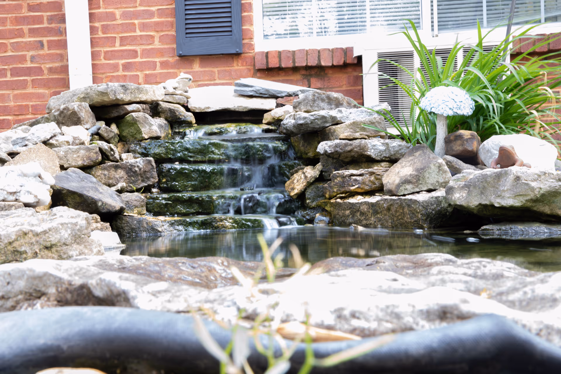 A small outdoor water feature with a cascading waterfall made of stacked rocks, surrounded by plants and a brick building wall with a window and air conditioning unit in the background.