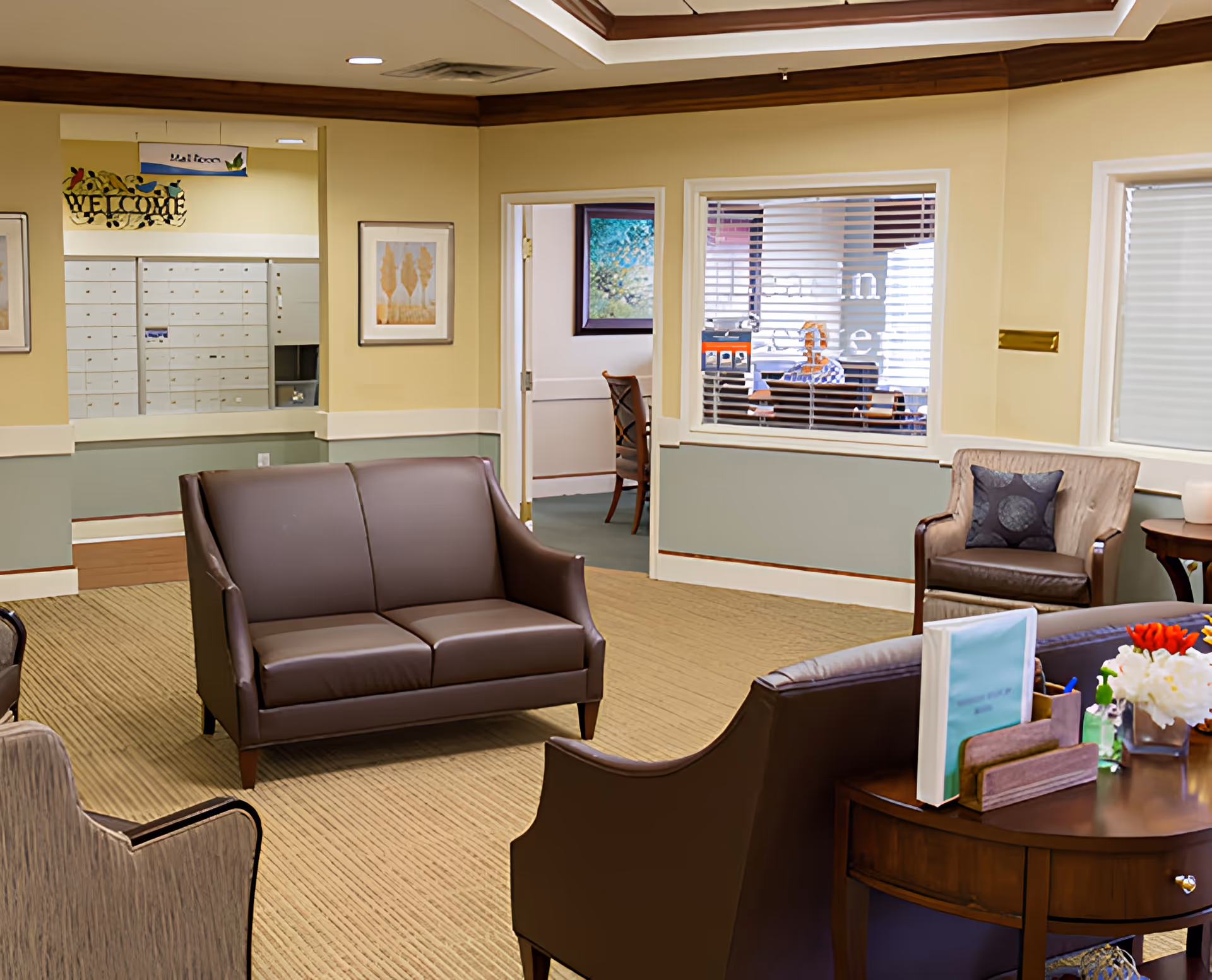 A cozy senior living facility common area with brown leather sofas and armchairs arranged on a beige carpet. There is a wooden table with flowers and documents in the foreground. The walls are painted yellow and green with framed artwork. A window with blinds reveals a room with a person sitting inside, and a mailroom with mailboxes is visible in the background.