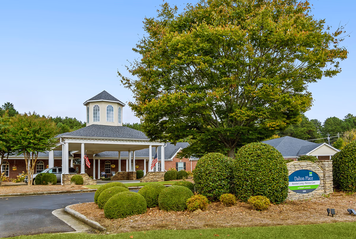 Exterior view of Dalton Place assisted living and memory care facility with a covered entrance, manicured bushes, trees, and a clear blue sky.