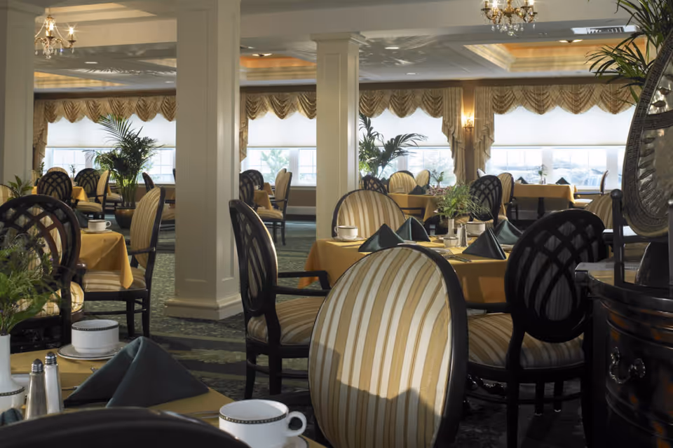 Elegant dining room with round tables covered in yellow tablecloths, set with white cups, saucers, and folded dark green napkins. The room features striped upholstered chairs, large windows with decorative valances, indoor plants, chandeliers, and white columns.