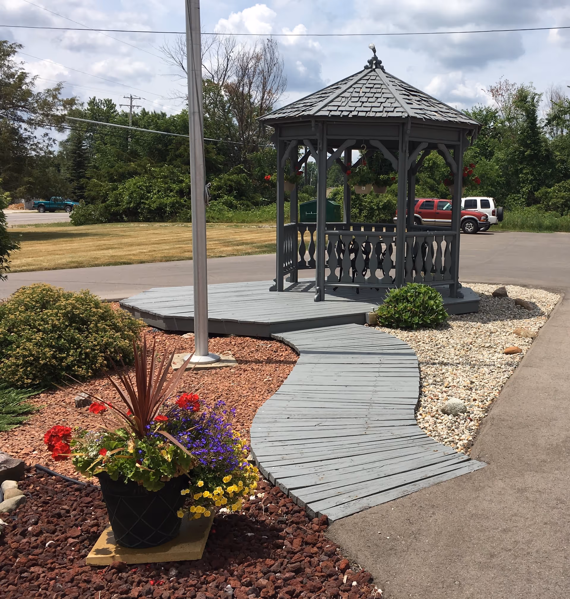 A gray wooden gazebo with a shingled roof sits at the end of a curved wooden walkway surrounded by landscaped areas with red mulch, rocks, and green bushes. A black planter with colorful flowers is in the foreground. In the background, there are trees, a grassy area, and parked vehicles under a partly cloudy sky.