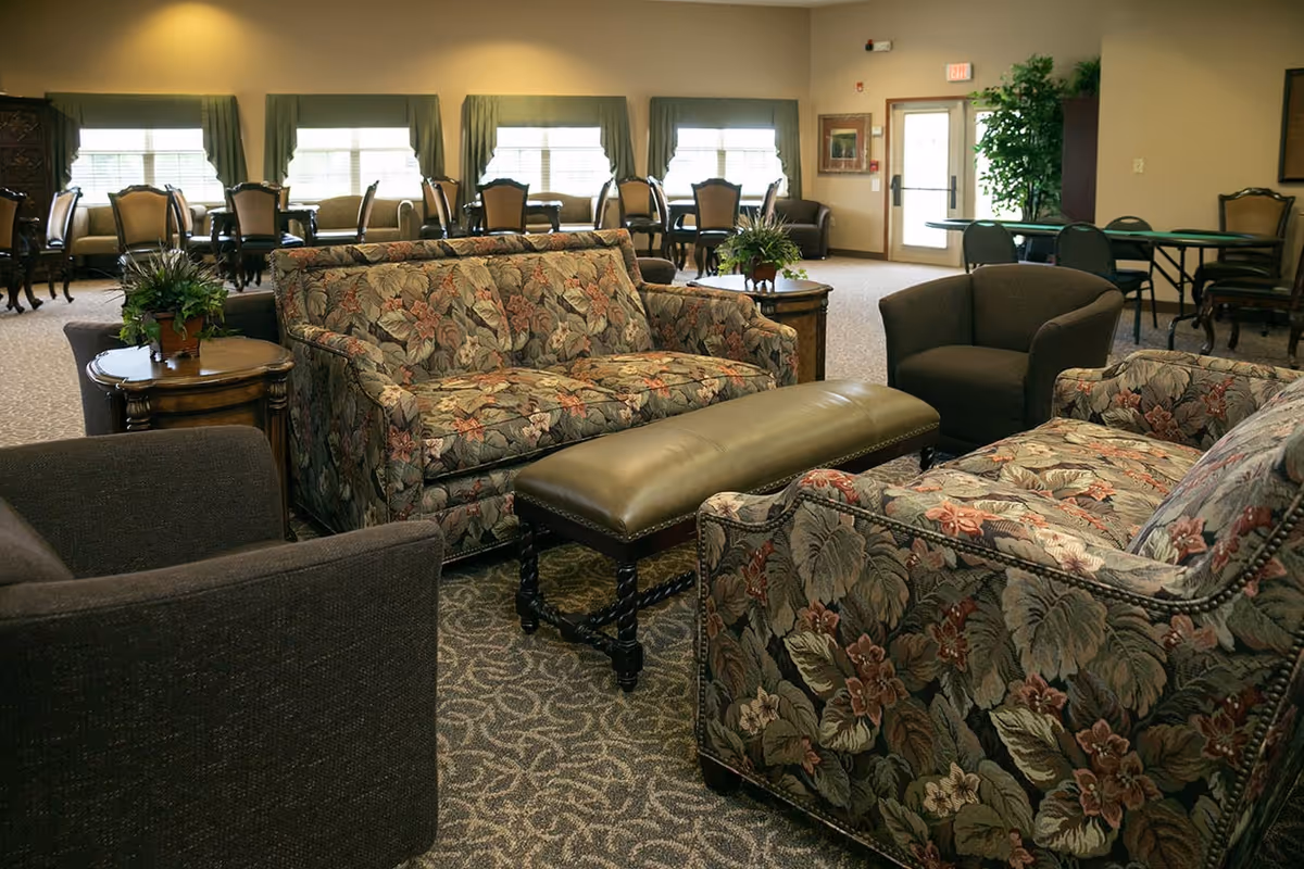 A spacious senior living common area with floral patterned sofas, dark armchairs, a long cushioned bench, and wooden side tables with potted plants. In the background, there are multiple dining tables and chairs near windows with green curtains, and a door leading outside.