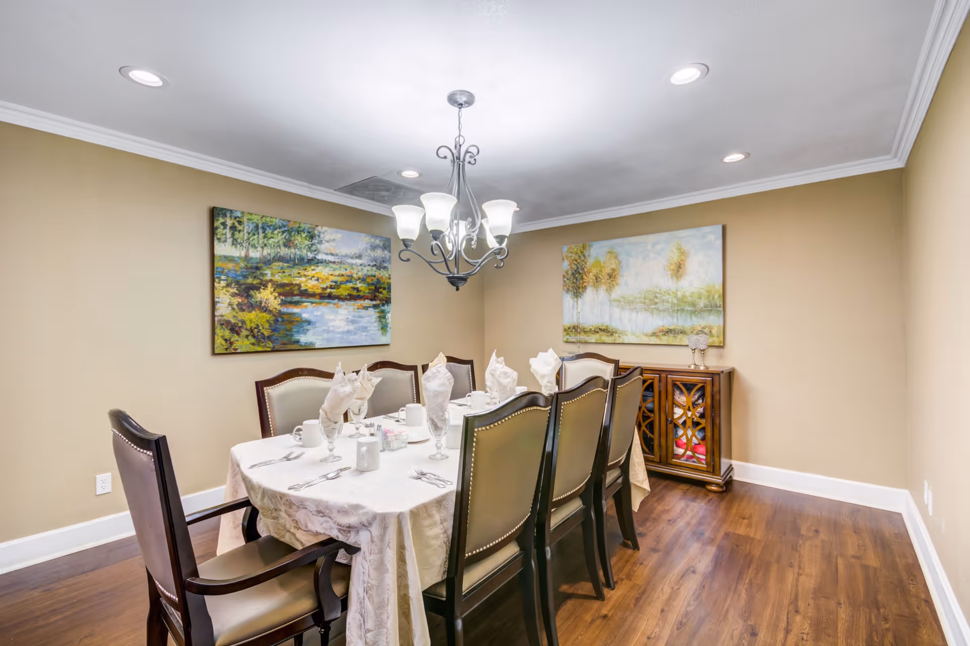 A formal dining room with a rectangular table covered with a white tablecloth. The table is set with folded napkins, glasses, mugs, and silverware. There are eight upholstered chairs around the table. The room has beige walls, wooden flooring, two landscape paintings on the walls, a wooden cabinet, and a chandelier hanging from the ceiling.