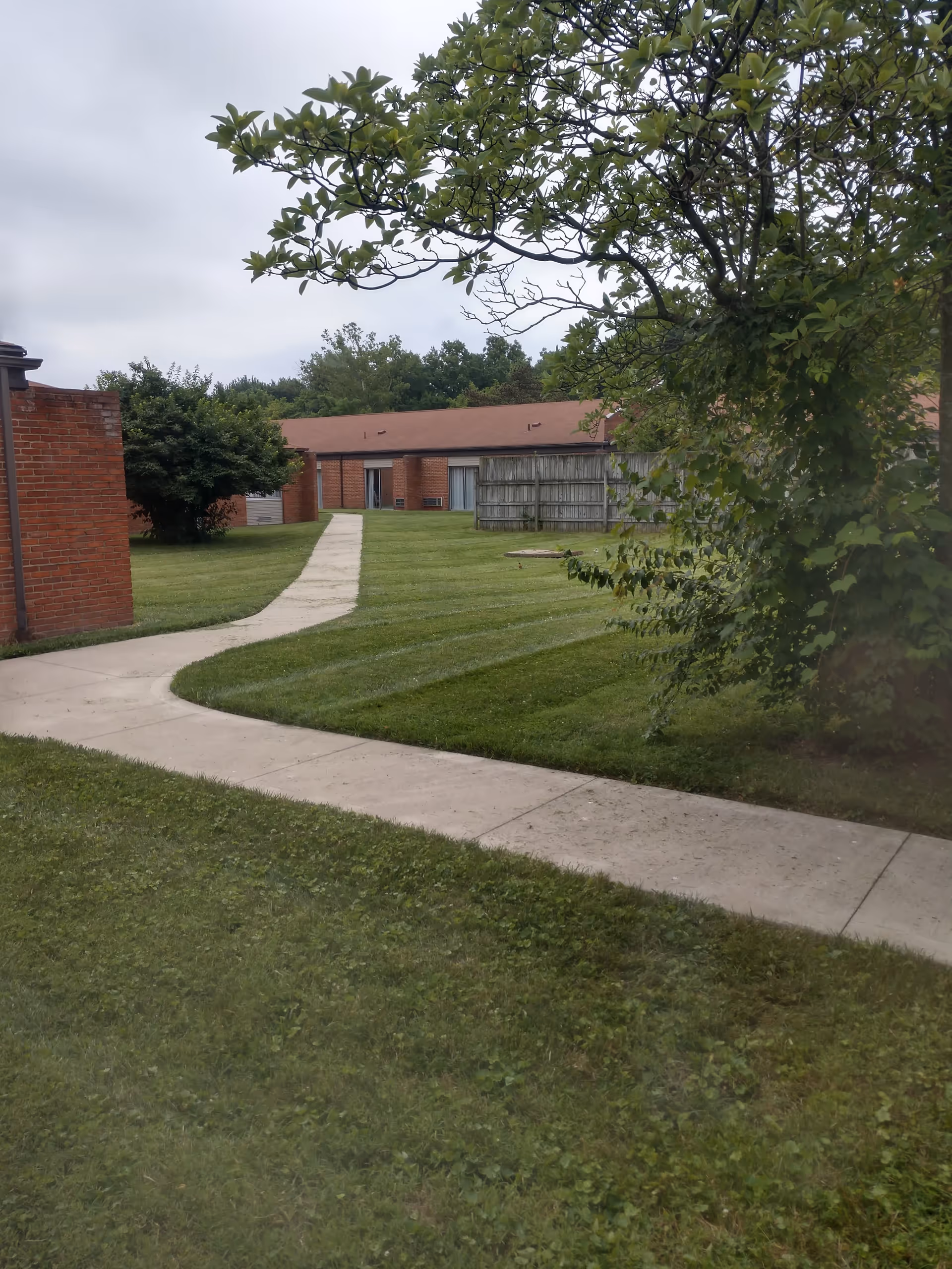 A paved walkway curves through a grassy outdoor area with trees and bushes, leading to a single-story brick building with a red roof in the background under a cloudy sky.