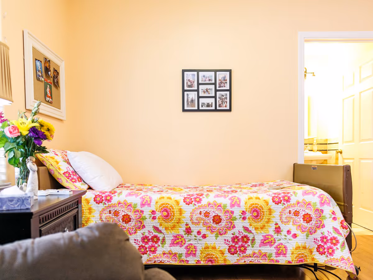 A cozy bedroom with a single bed covered in a colorful floral quilt in shades of pink, yellow, and orange. There is a white pillow and a floral pillow on the bed. To the left of the bed is a dark wooden nightstand with a lamp, a vase of fresh flowers, and some small decorative items. On the wall above the bed is a collage picture frame with multiple photos. An open door on the right side reveals a glimpse of a bathroom with a sink and mirror.