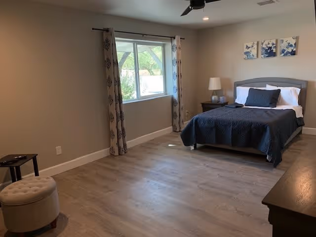 Spacious bedroom with a bed, nightstand and lamp near a window with patterned curtains, wood-look flooring and neutral walls.