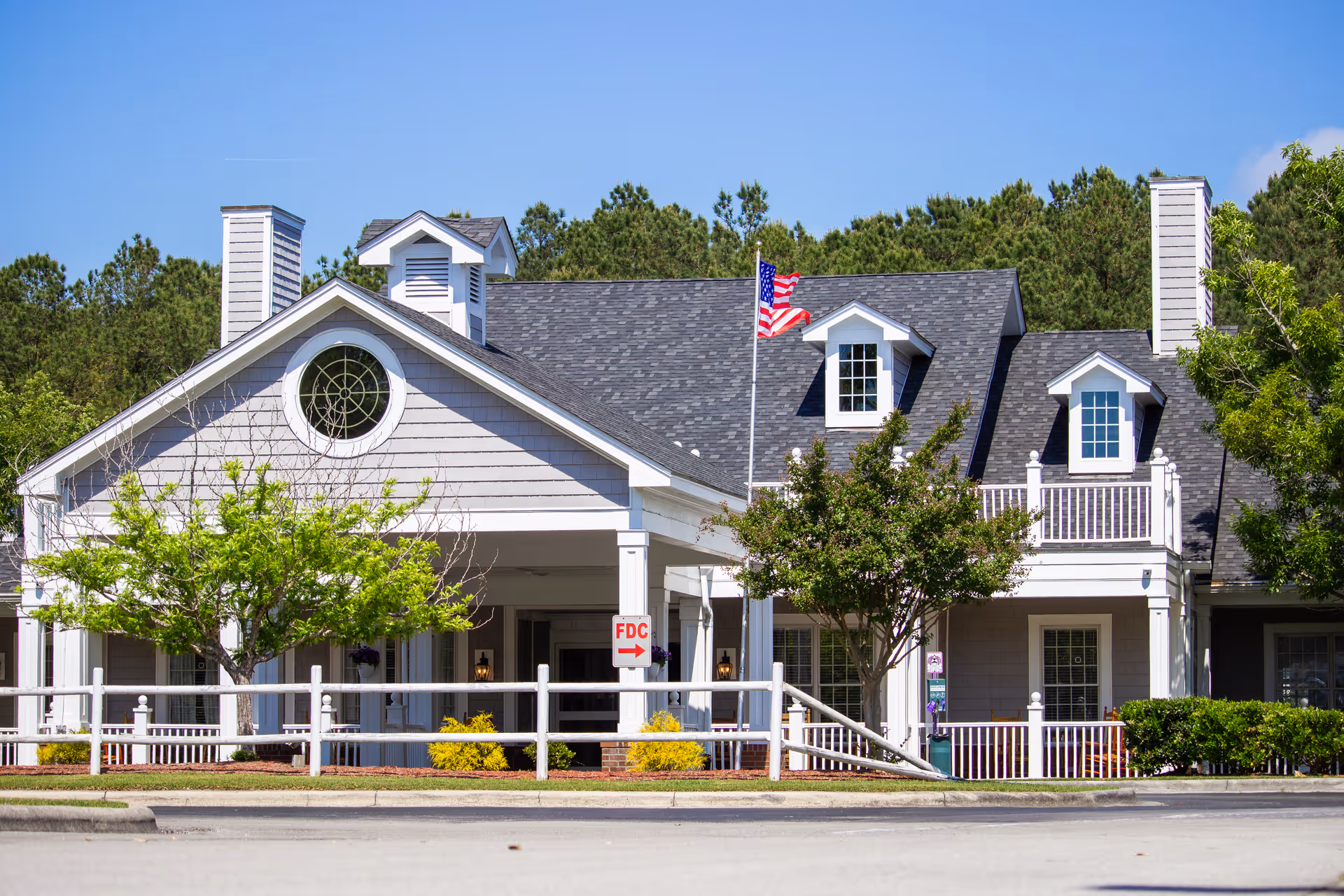 Front exterior of a two-story senior living building with white siding, dormer windows, an American flag, porch and landscaped trees.