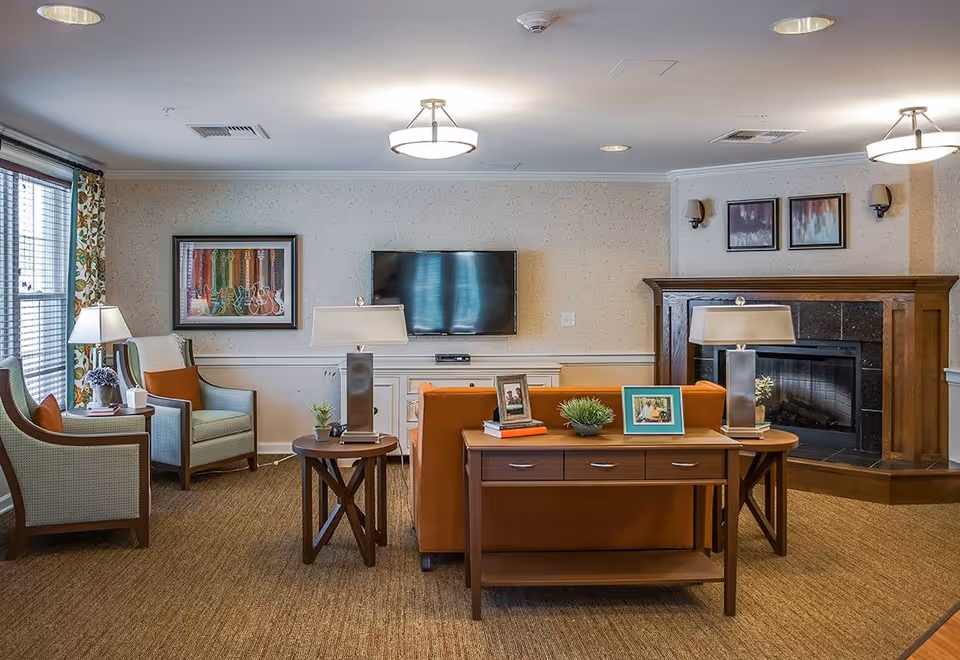 A cozy living room in a senior living facility featuring a brown sofa with a wooden console table behind it, two armchairs with orange cushions near a window with floral curtains, a flat-screen TV mounted on the wall, a fireplace with a wooden mantel, and several framed artworks on the walls. The room is warmly lit with ceiling lights and table lamps.