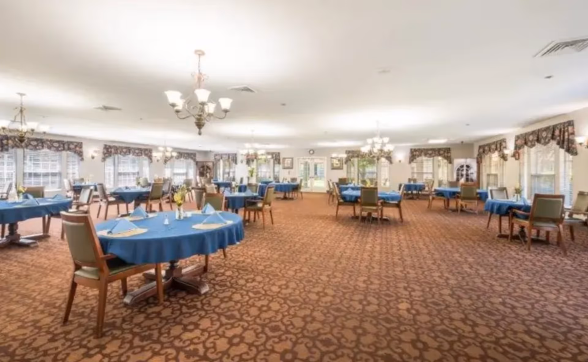 Spacious dining room with round tables covered in blue tablecloths, dining chairs, chandeliers, and patterned carpet.
