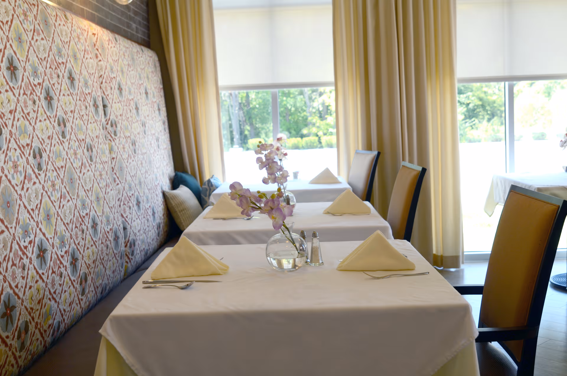Sunlit dining room with white-tablecloth tables set with folded napkins, a small vase of flowers, and patterned banquette seating by windows.