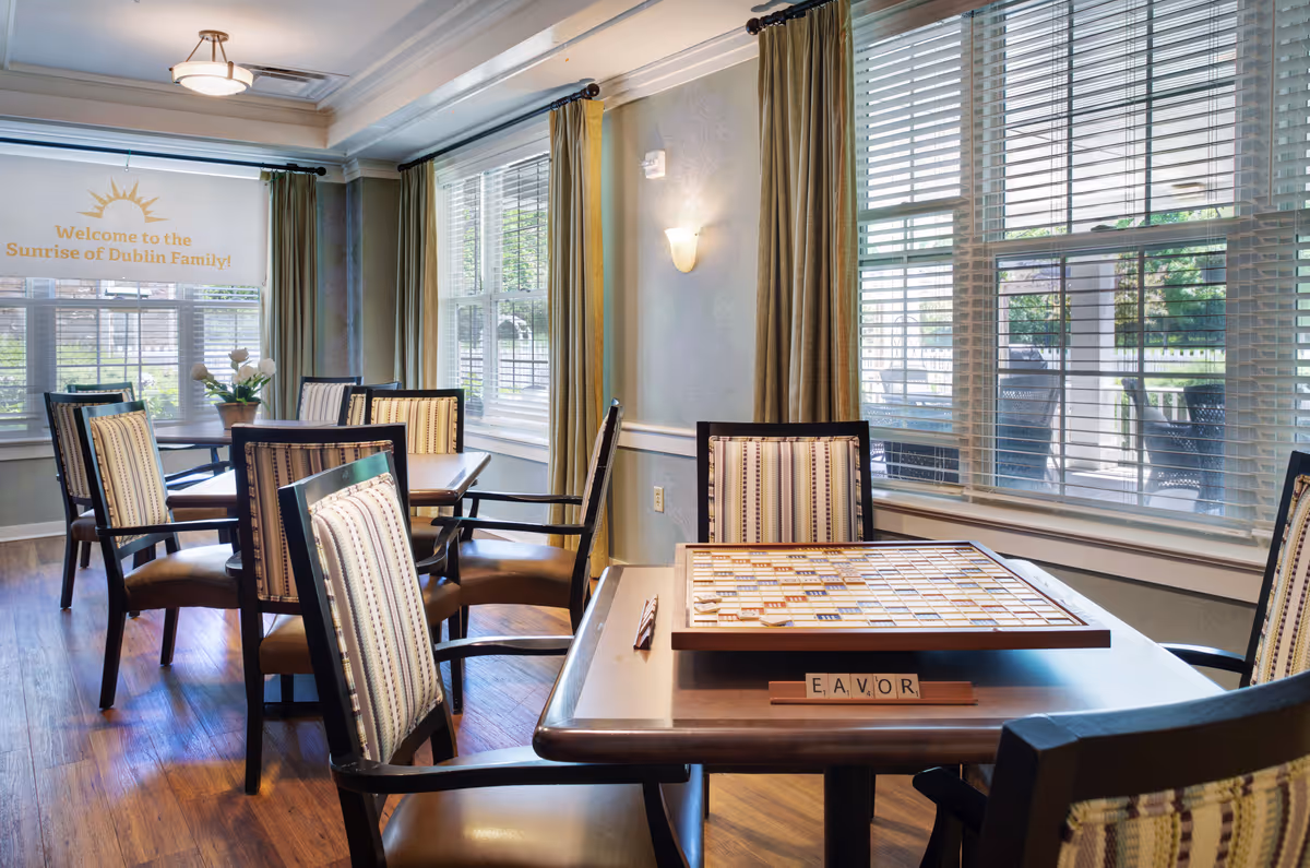 A bright and inviting common area in Sunrise of Dublin with several wooden tables and cushioned chairs arranged neatly. One table in the foreground has a Scrabble board game set up with letter tiles spelling 'EAVOR'. Large windows with blinds and curtains allow natural light to fill the room. A banner on the window reads 'Welcome to the Sunrise of Dublin Family!'.