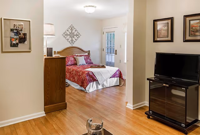 A cozy bedroom with a wooden bed frame, red floral bedspread, and a white throw blanket. The room has hardwood floors, a wooden dresser with a lamp, and a wall decoration above the bed. A TV is placed on a black stand against the wall, with two framed pictures hanging above it. There is a door with glass panels leading outside or to another room.