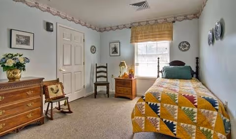 Small traditional bedroom with a single bed dressed in a colorful patchwork quilt, wooden dresser, nightstand, chair, and a window with a valance.