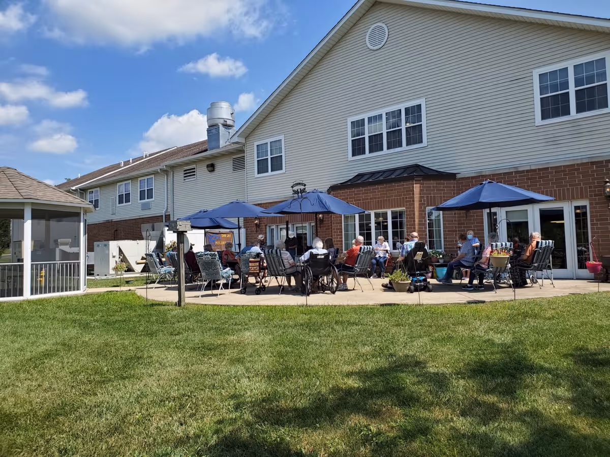 Outdoor patio area at a senior living facility with several elderly people sitting on chairs under blue umbrellas, enjoying the sunny day. The building has beige siding with brick accents and multiple windows. There is a grassy lawn in the foreground and a gazebo on the left side.