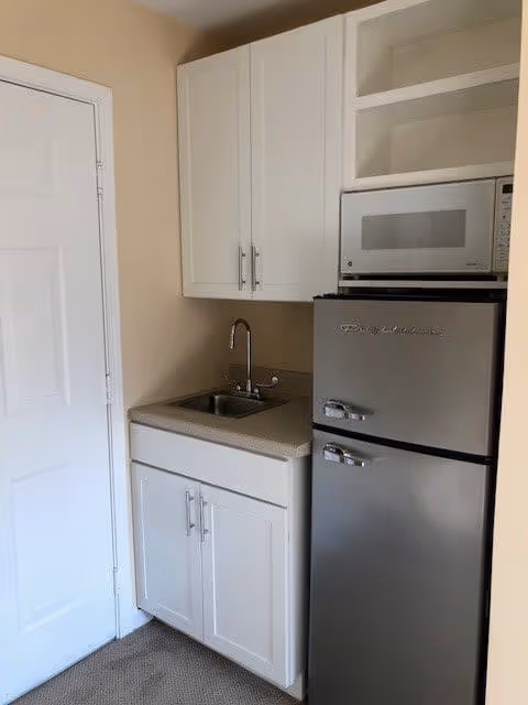 Small kitchenette area with white cabinets, a stainless steel mini refrigerator, a microwave oven on top of the refrigerator, a countertop with a small sink and faucet, and a closed white door to the left.