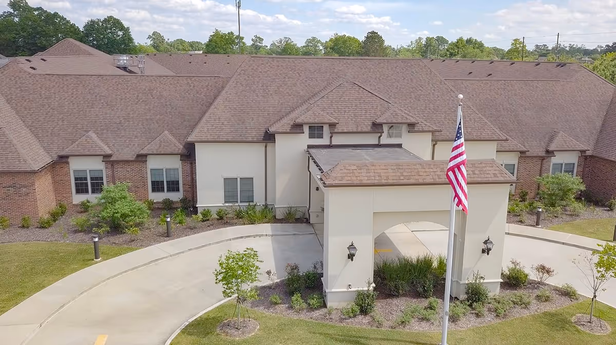 Exterior view of a single-story senior living facility building with a brown shingle roof and beige walls. The entrance features a covered driveway with two wall-mounted lanterns and an American flag on a flagpole in front. The surrounding area has landscaped greenery and a curved driveway.