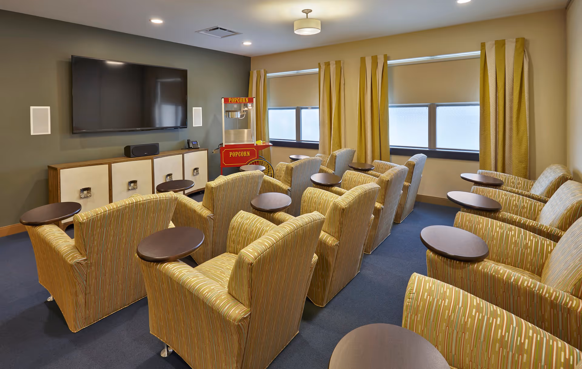 A small theater room with multiple rows of cushioned armchairs, each equipped with a small attached table. A large flat-screen TV is mounted on the wall above a cabinet. There is a red popcorn machine in the corner near windows with yellow curtains.