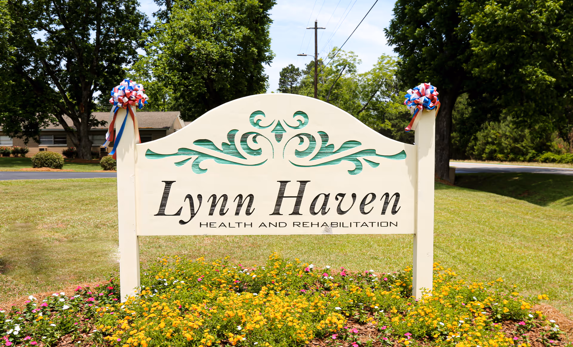 A white wooden sign with green decorative accents that reads 'Lynn Haven Health and Rehabilitation' surrounded by a flower bed with yellow and pink flowers, set on a grassy area with trees and a road in the background.