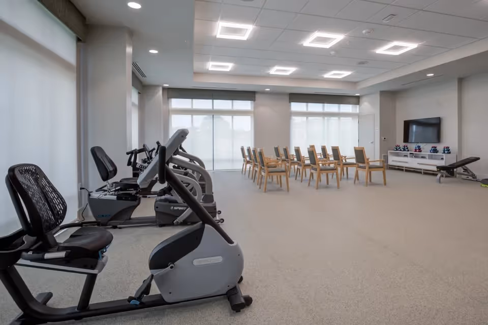 A spacious fitness and activity room with exercise equipment including stationary bikes on the left side and a row of wooden chairs arranged in the center facing a wall-mounted TV. The room has large windows with blinds, neutral-colored walls, and modern ceiling lights.