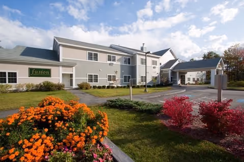 Exterior view of a two-story senior living facility building with a covered entrance. The building is surrounded by a well-maintained lawn, colorful flower beds with orange and red flowers, and a clear blue sky with some clouds.