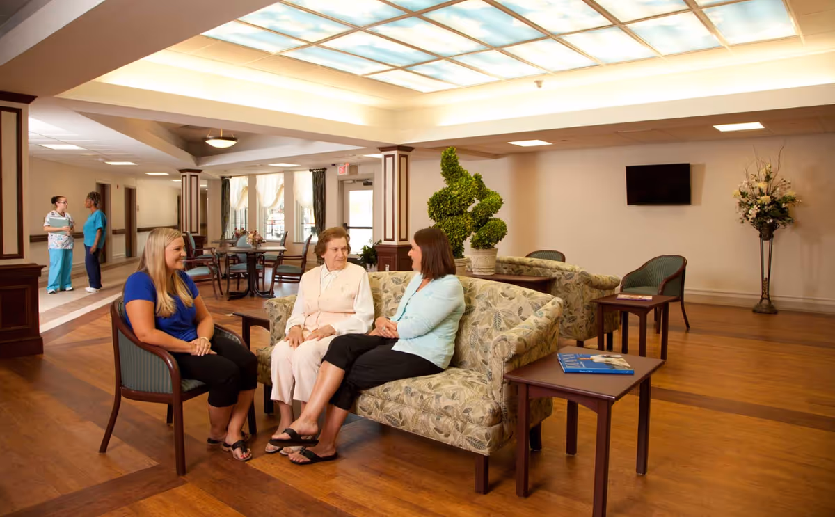 A well-lit common area in an assisted living facility with three women sitting and conversing on floral-patterned sofas and chairs. In the background, two staff members are talking near a hallway. The room has wooden flooring, a large ceiling light panel, a TV mounted on the wall, and decorative plants and flowers.