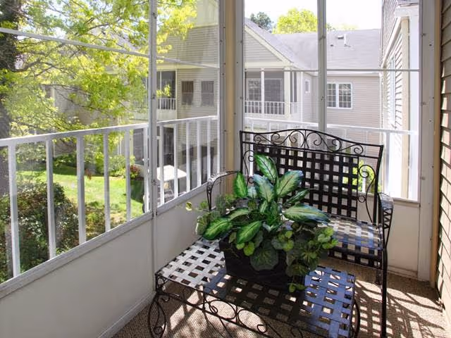 A screened-in balcony with a black metal bench and matching table, both featuring a lattice design. A potted green plant sits on the table. The balcony overlooks a grassy area with trees and neighboring buildings in the background.