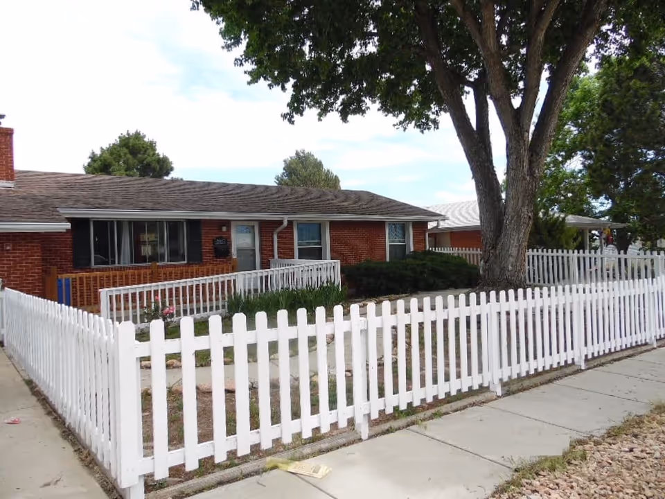 Single-story brick building with a white picket fence surrounding a small yard with a large tree and some bushes, under a partly cloudy sky.
