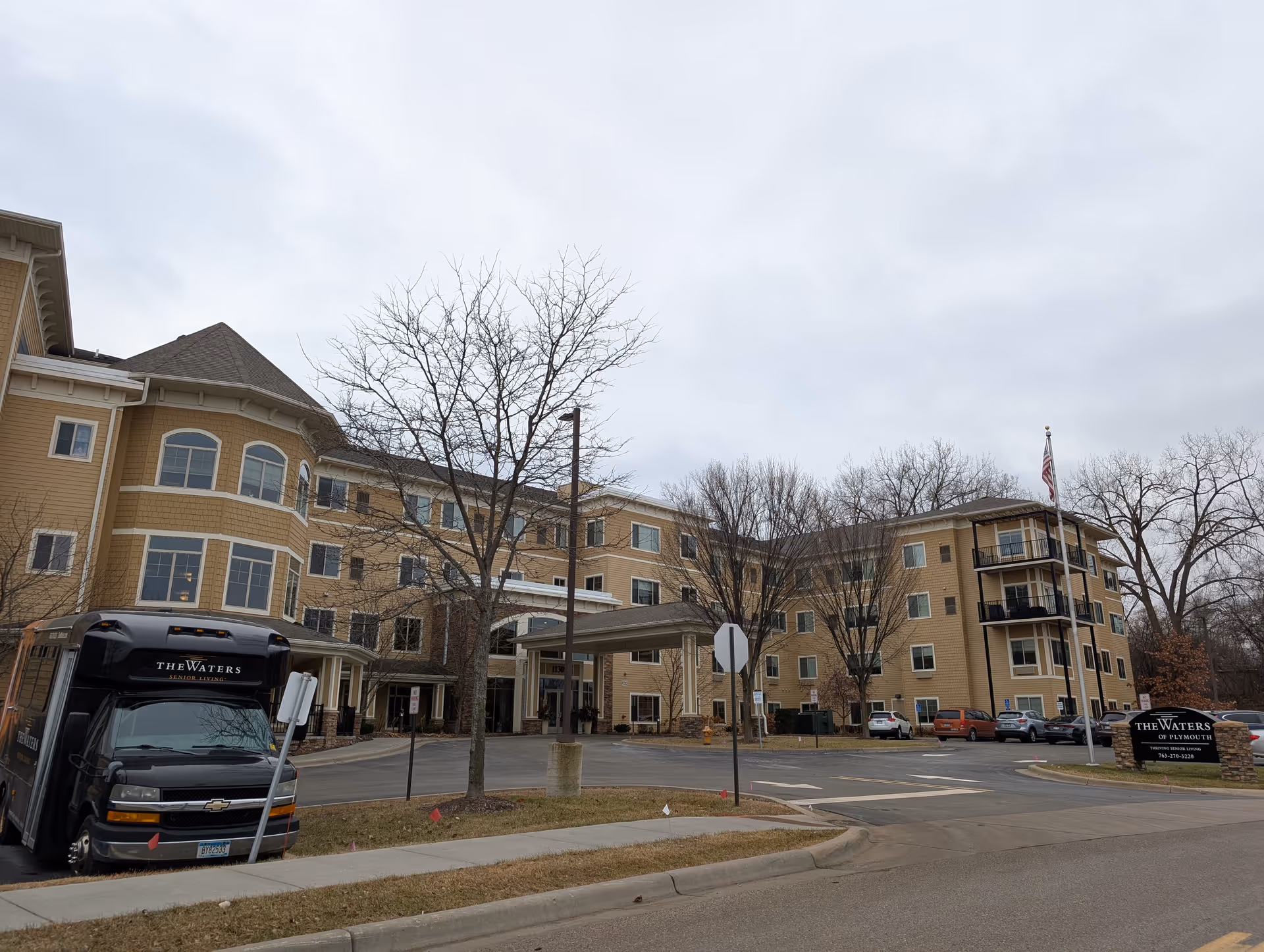 Exterior view of The Waters of Plymouth senior living facility showing a large multi-story beige building with multiple windows, a covered entrance, a parking area with several cars, a black shuttle bus with The Waters Senior Living logo, leafless trees, and an American flag on a flagpole.