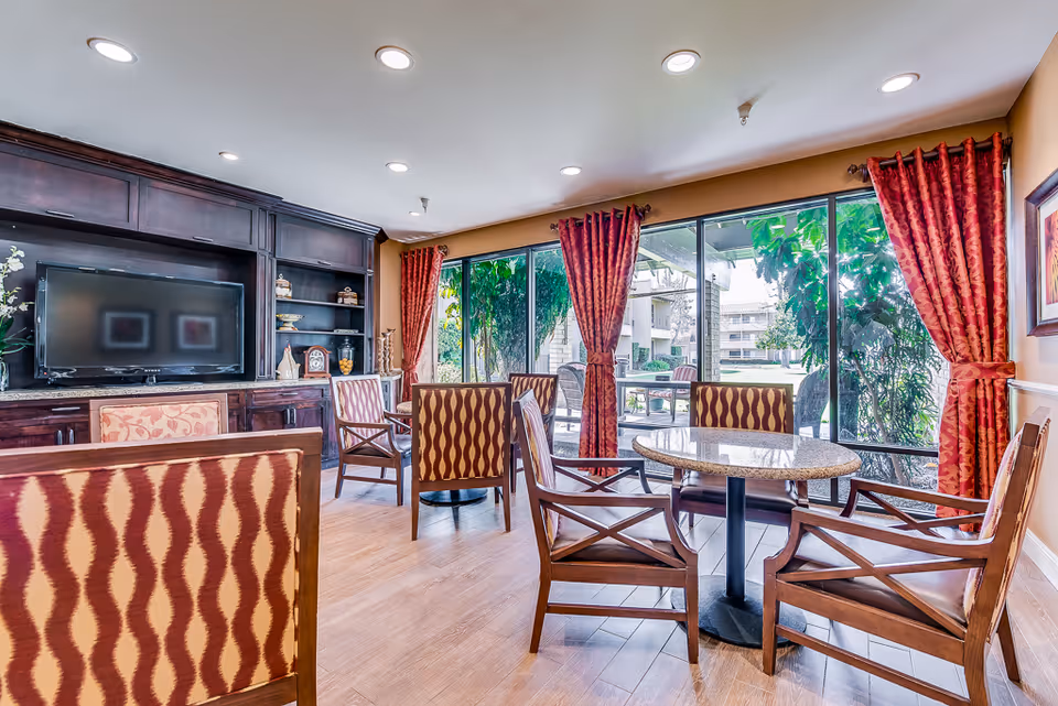 Bright communal dining/lounge area with round tables, patterned chairs, a built-in TV cabinet, and large windows with red curtains overlooking greenery.