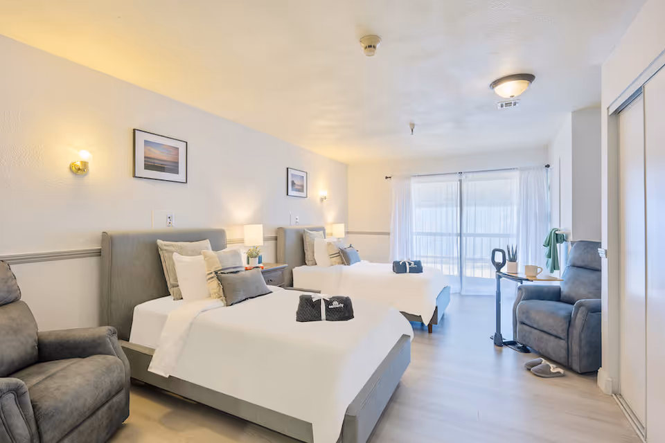 A bright and spacious bedroom with two single beds, each with white bedding and multiple pillows. There are two bedside tables with lamps and small plants. The room features two comfortable gray armchairs, a small table with a cup and a plant, and large windows with sheer white curtains allowing natural light to fill the space. The walls are decorated with framed pictures and soft wall lights.