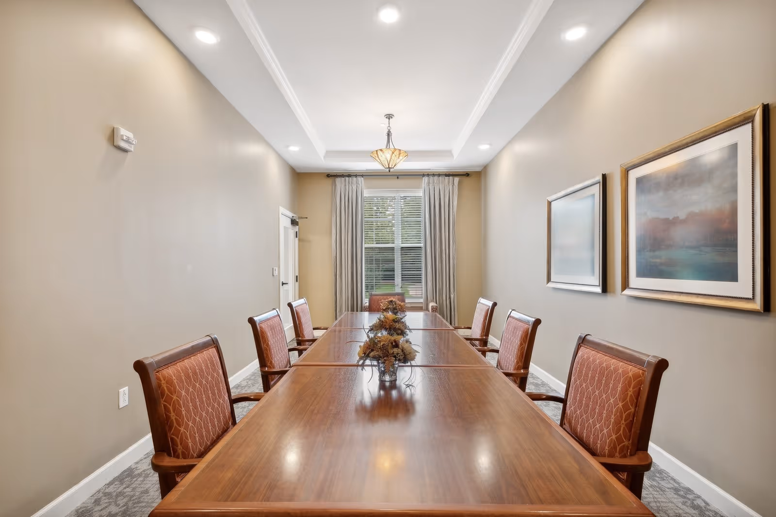A formal dining room with a long wooden table surrounded by eight upholstered chairs with wooden frames. The room has beige walls, two framed paintings on the right wall, a window with curtains at the far end, and a ceiling light fixture above the table.