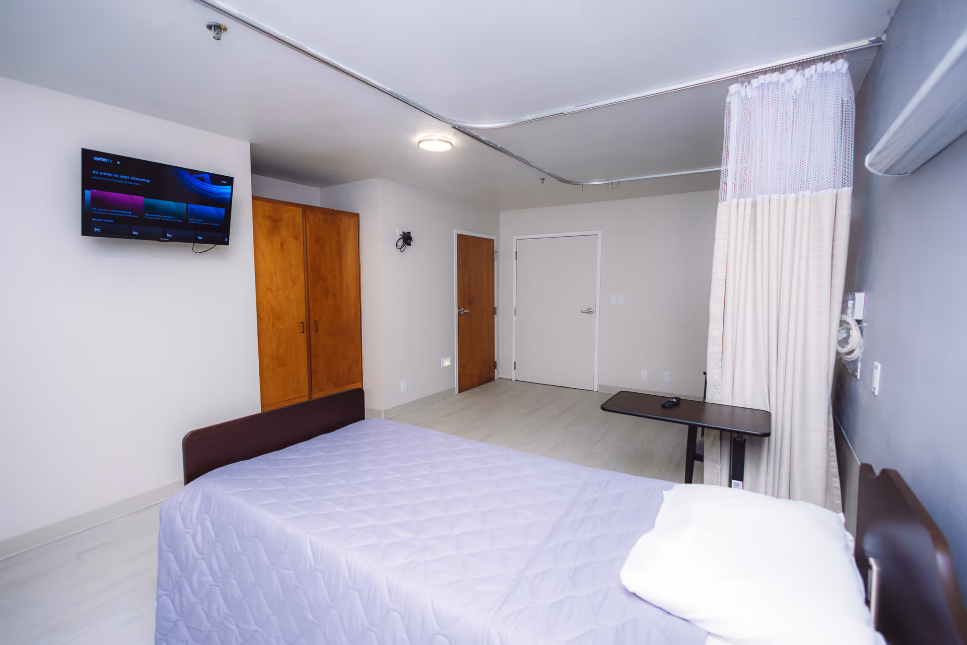 A simple, clean bedroom in a healthcare facility with a single bed covered in a light purple quilt, a pillow, a wall-mounted TV, wooden closet doors, a small overbed table, and a privacy curtain partially drawn.