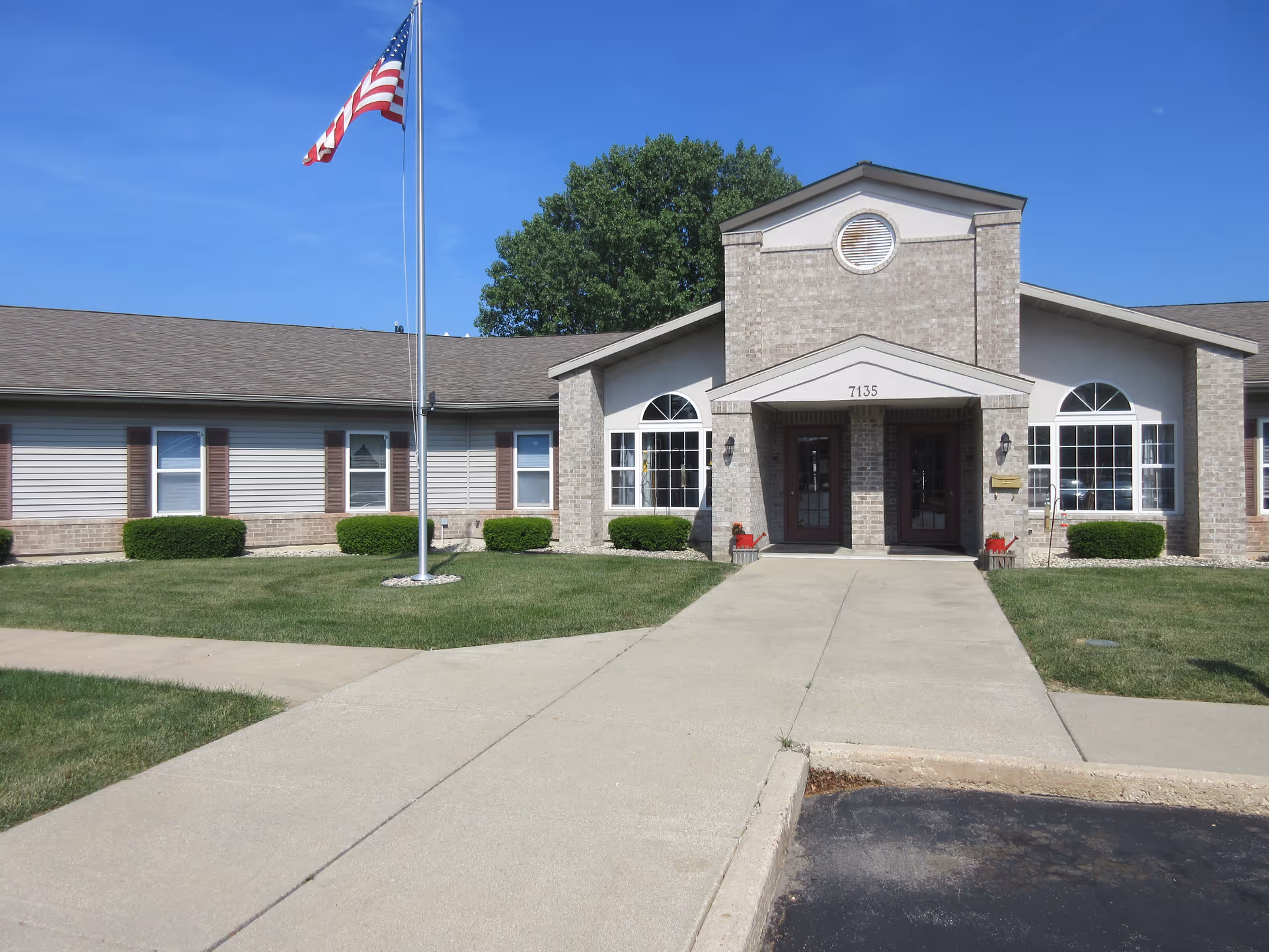 Front entrance of a single-story brick and siding building with a flagpole, concrete walkway, and manicured lawn.