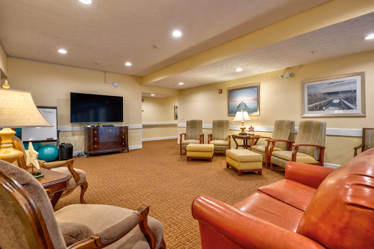 A cozy living room area with a red leather sofa, several upholstered armchairs with matching ottomans, a wooden side table with a lamp, a flat-screen TV mounted on a wooden cabinet, and framed pictures on the walls. The room has beige walls, carpeted flooring, and recessed ceiling lights.