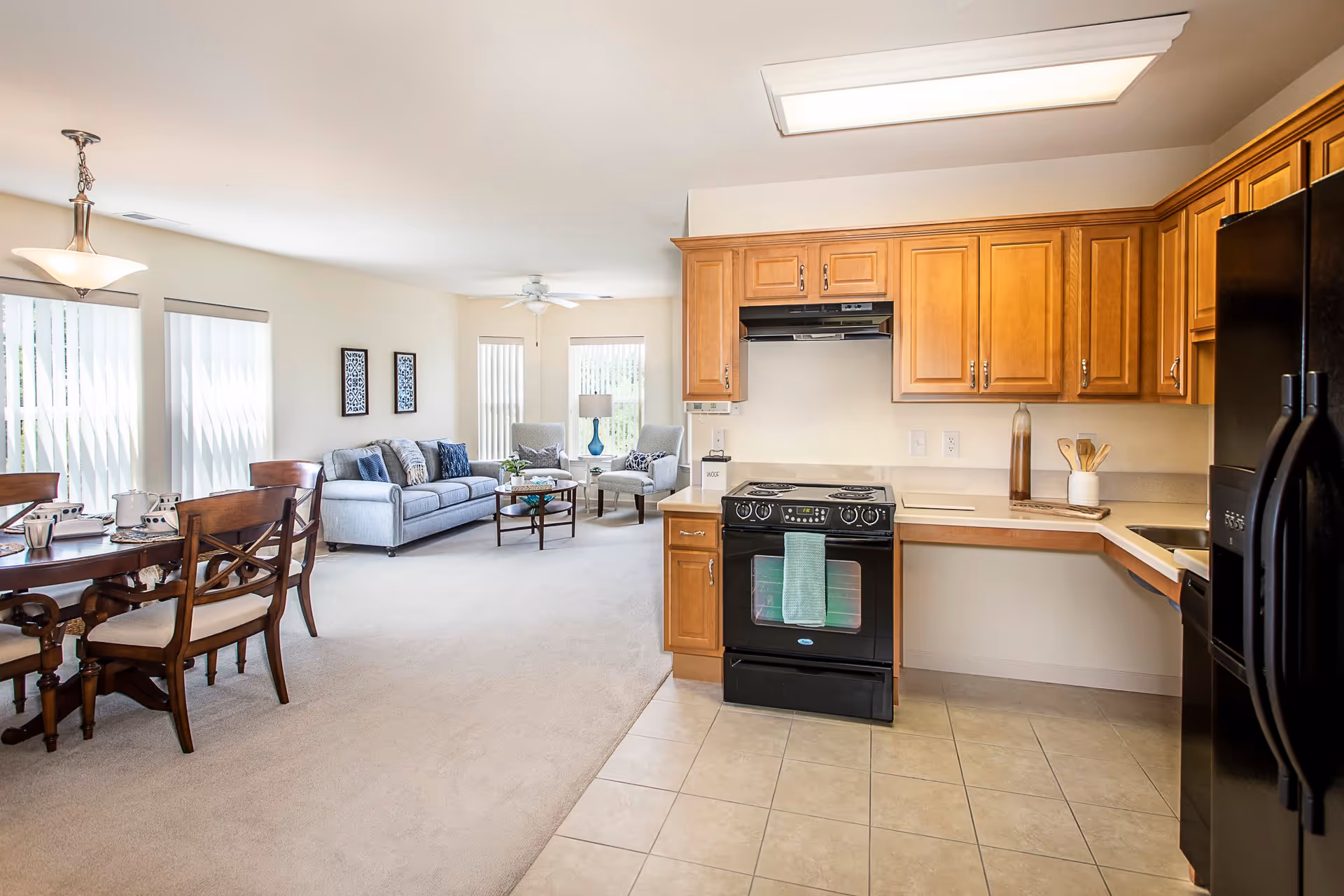 Open concept living space featuring a kitchen with wooden cabinets, black appliances including a stove and refrigerator, and a dining area with a wooden table and chairs. The living room area has a gray sofa, two armchairs, a coffee table, and large windows with vertical blinds allowing natural light to fill the room.