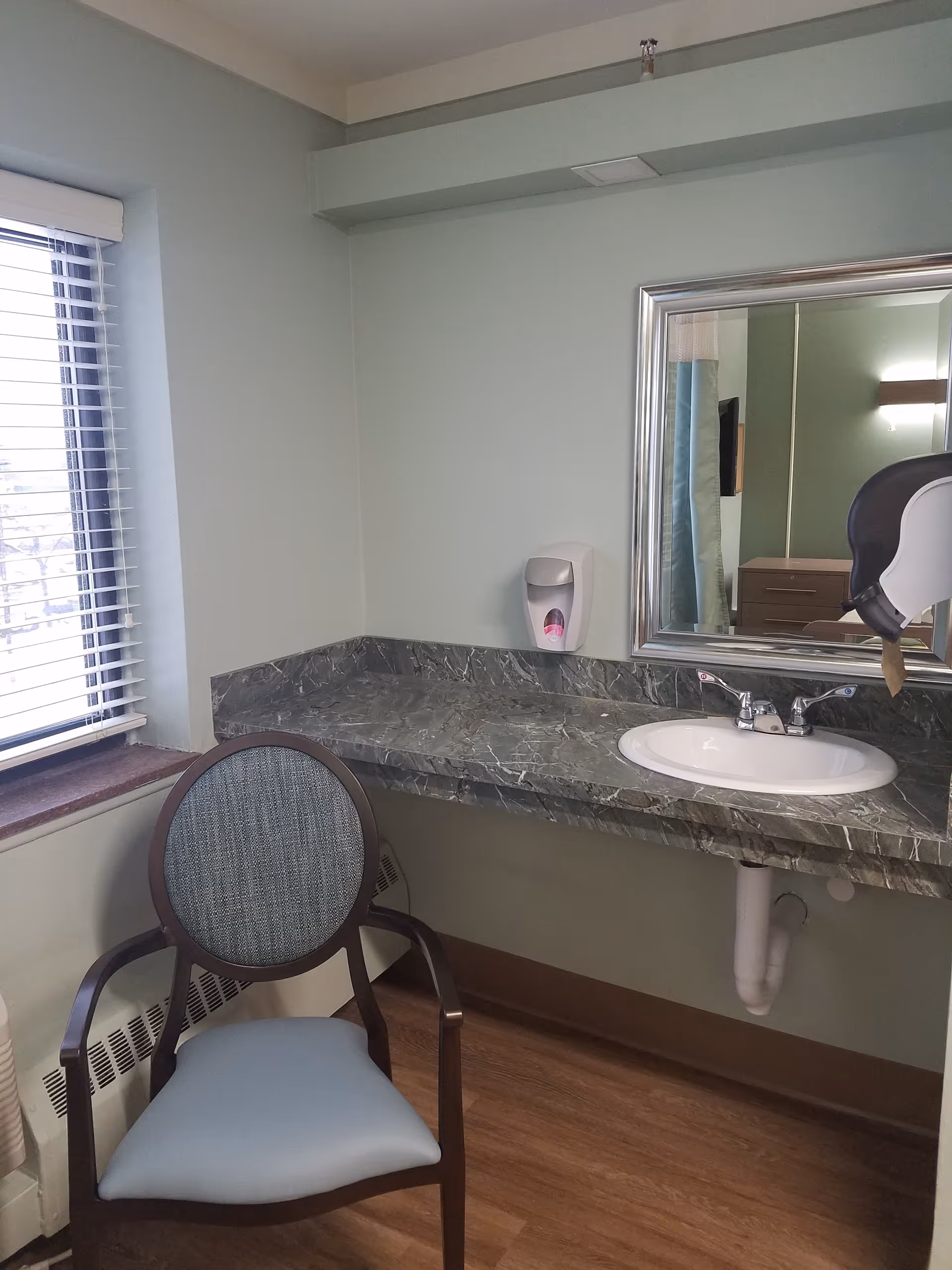 A small room corner with a green marble countertop featuring a white sink and a silver-framed mirror above it. A soap dispenser is mounted on the wall next to the mirror. In front of the countertop, there is a wooden chair with a blue cushioned seat and backrest. A window with white blinds is on the left side, and the floor is wooden.