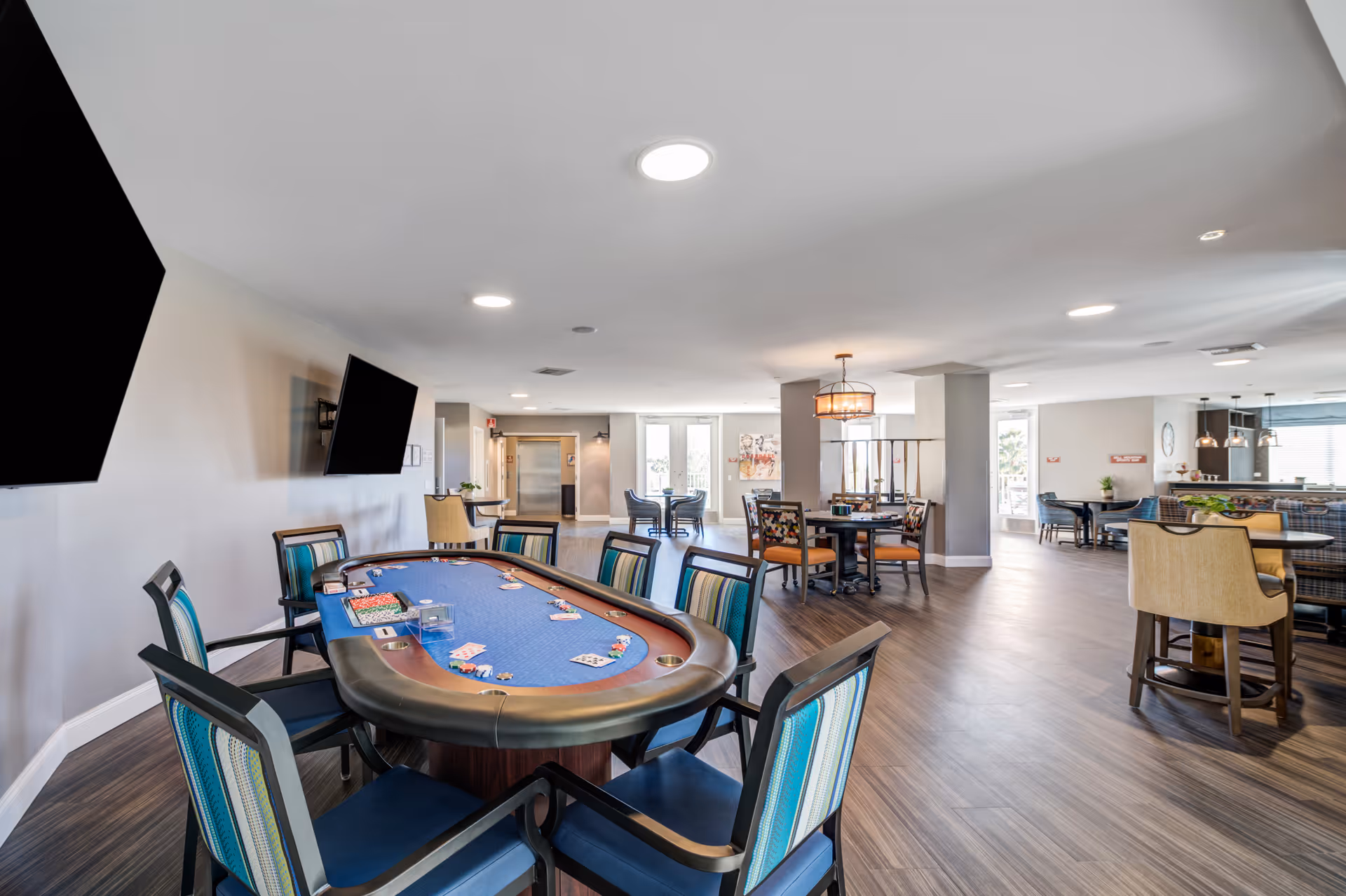 A spacious and well-lit common area in a senior living facility featuring a poker table with playing cards and chips, surrounded by chairs with blue and striped upholstery. The room has multiple seating areas with tables and chairs, flat-screen TVs mounted on the wall, and large windows allowing natural light to enter. The flooring is wood-style, and the ceiling has recessed lighting.