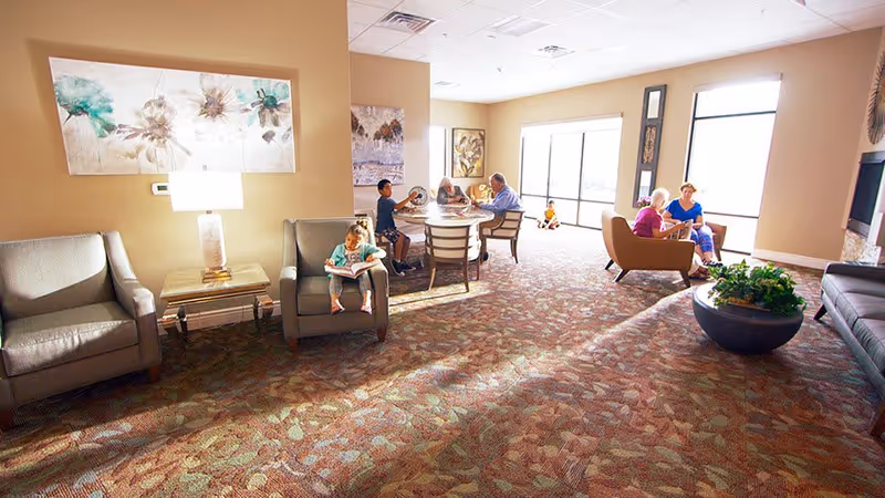 A bright and spacious living room area in an assisted living center with several people sitting and engaging in activities. There are armchairs, a round table with chairs, large windows letting in natural light, and decorative artwork on the walls. A child is sitting on an armchair reading a book, while other individuals are seated around the table and in chairs near the windows.