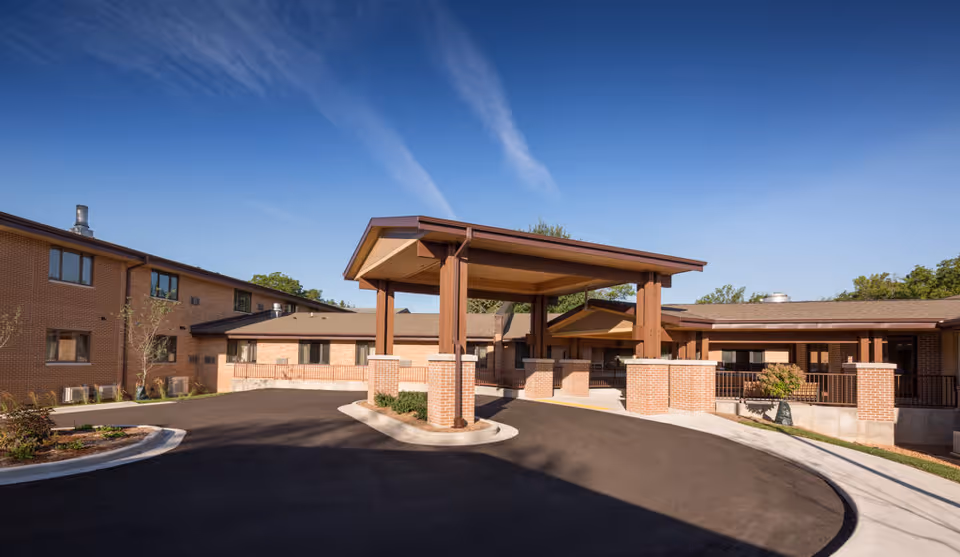 Front entrance of a brick senior living building with a covered porte-cochere and circular driveway under a clear blue sky.