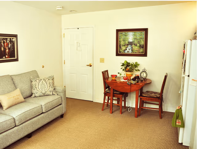 A cozy interior room featuring a gray upholstered sofa with decorative pillows on the left, a small wooden dining table with two chairs against the wall, a potted plant, and a clock on the table. A framed painting of a forest and stream hangs above the table. A white door is centered in the background, and a white refrigerator is visible on the right side of the image.
