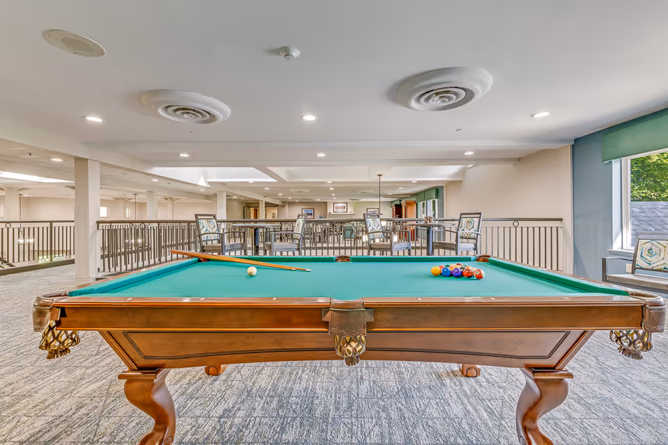 A pool table set up in a spacious, well-lit common area with seating and windows.