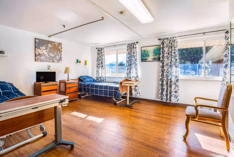 A bright and clean room with two single beds covered in blue checkered bedding, a wooden floor, a small TV on a wooden dresser, a lamp, a wooden chair, and windows with floral curtains letting in natural light.