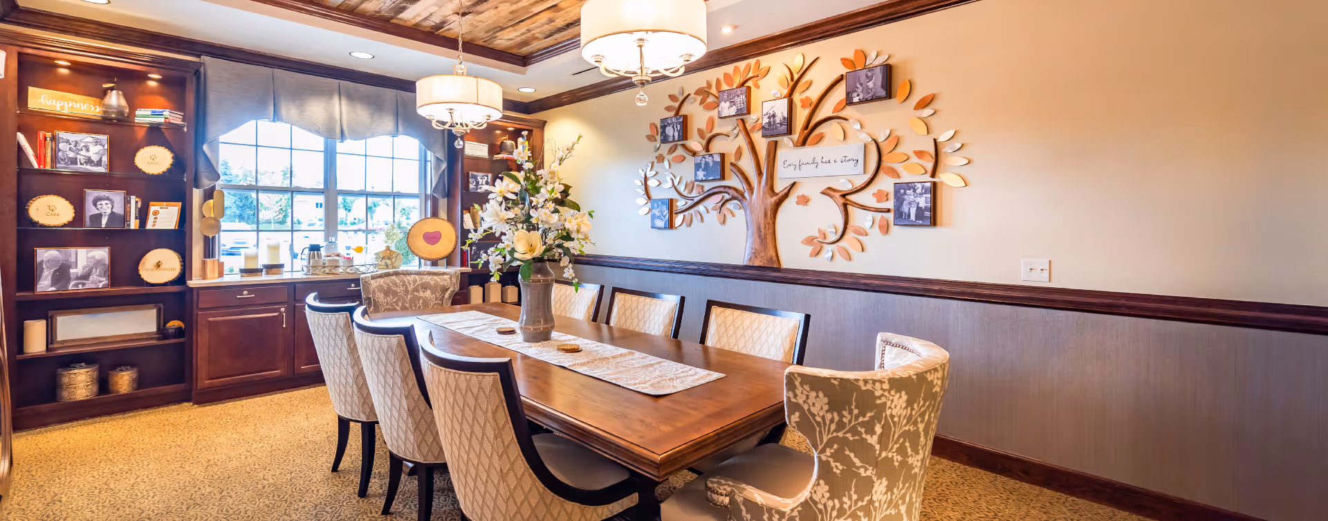 A warmly decorated dining room with a long wooden table surrounded by upholstered chairs. A vase with flowers is centered on the table. The room features a large window with a valance, a wooden bookshelf with framed photos and decorative items, and a wall decoration shaped like a tree with photo frames and a sign that reads 'Our family has a story.' The ceiling has wooden paneling and two hanging light fixtures.