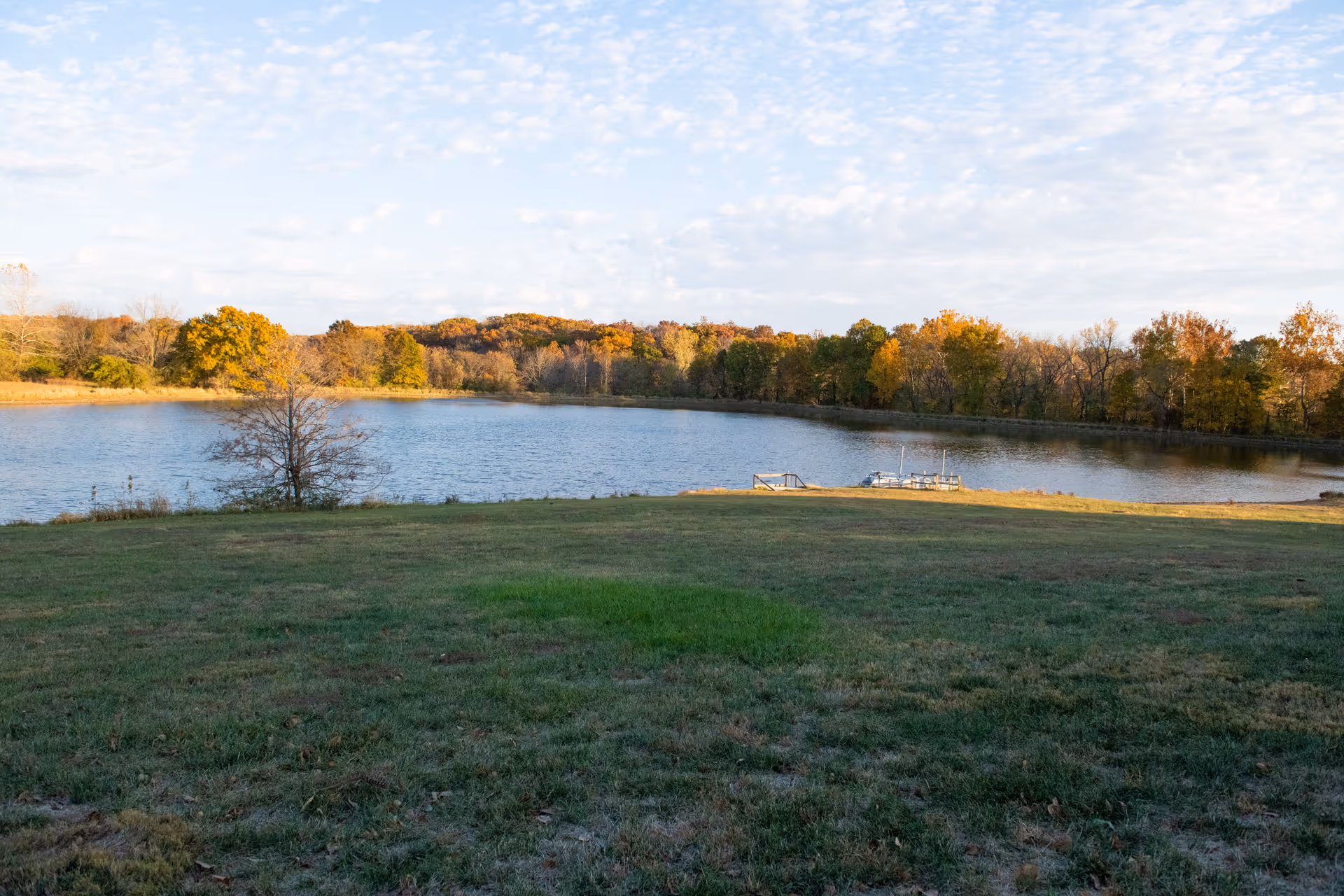 A scenic outdoor view of a lake surrounded by trees with autumn foliage, a grassy area in the foreground, and a small dock extending into the water under a partly cloudy sky.