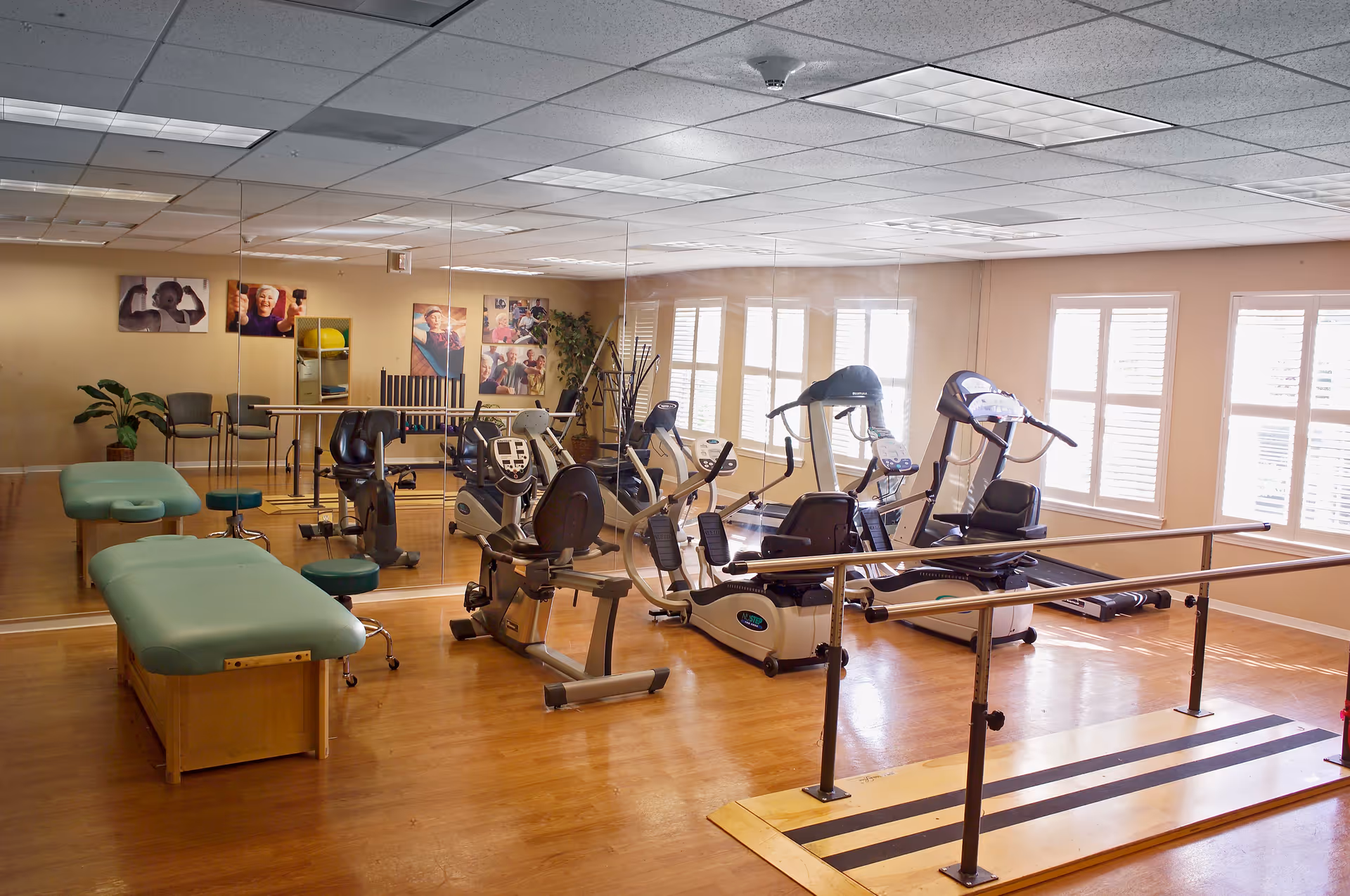 A bright exercise room with wooden floors featuring various fitness equipment including stationary bikes, treadmills, parallel bars for walking support, and padded therapy tables. The room has large windows with white shutters allowing natural light, a mirrored wall reflecting the equipment, and motivational photos on the far wall.