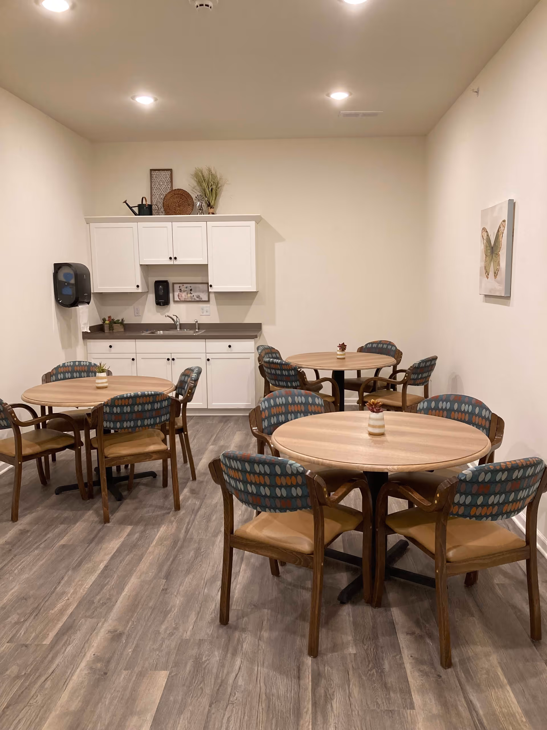 A small dining area with three round wooden tables, each surrounded by four chairs with patterned backs and tan seats. In the background, there is a kitchenette with white cabinets, a countertop, a sink, and a wall-mounted soap dispenser. The room has light-colored walls, wood-look flooring, and a framed butterfly picture on the right wall.