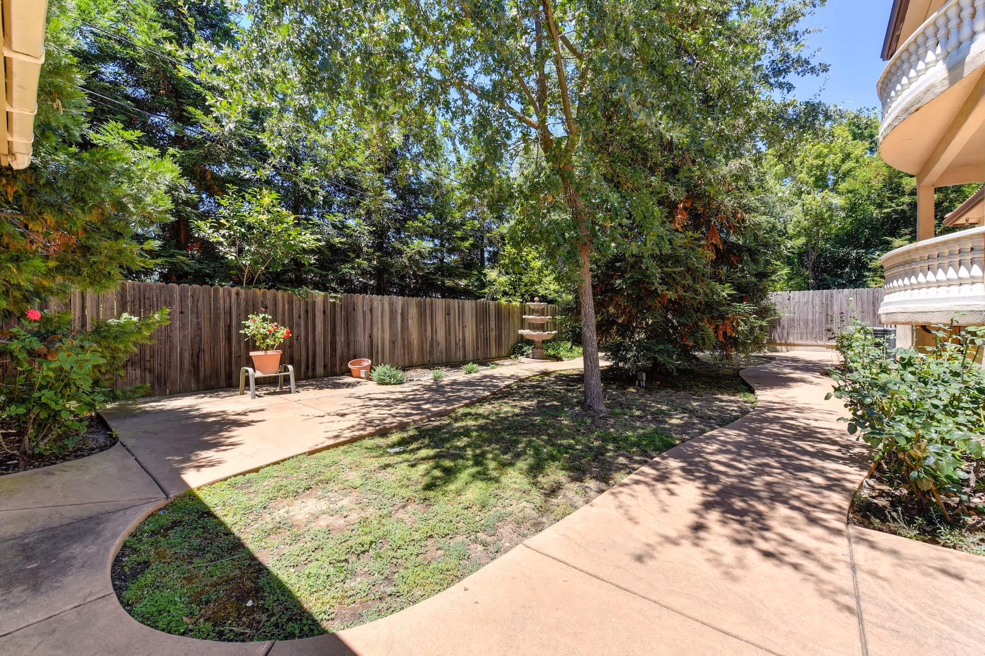 Outdoor garden area with a curved concrete pathway, green grass, trees, and potted plants along a wooden fence. Part of a building with balconies is visible on the right side.