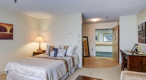 A well-lit bedroom in a senior living facility featuring a neatly made bed with multiple pillows, a bedside table with a lamp, a wooden desk with a chair, and framed artwork on the walls. The room has a doorway leading to a hallway with additional framed pictures visible.