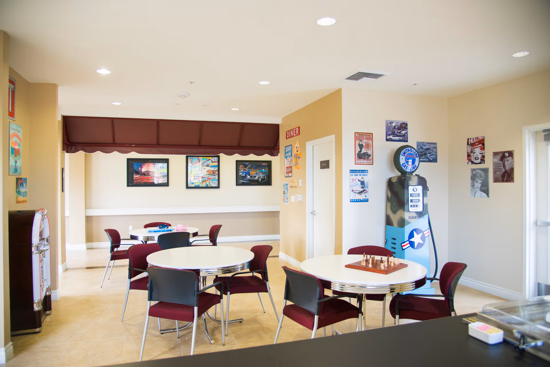 A bright and clean common area with round white tables and maroon chairs arranged for seating. The walls are decorated with vintage-style posters and artwork, including a retro gas pump and a small chessboard on one table. The room has beige tiled floors and recessed ceiling lights, creating a welcoming atmosphere.