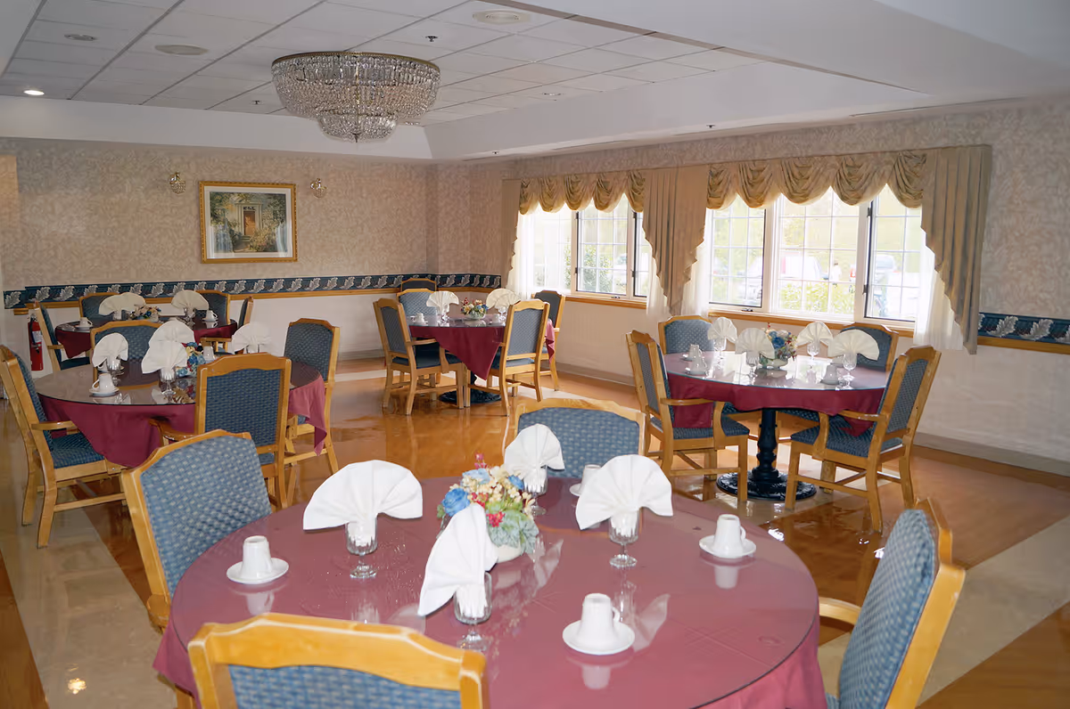 A dining room with round tables covered in maroon tablecloths, each set with folded white napkins, cups, and glasses. The room has wooden chairs with blue cushions, large windows with beige curtains, a chandelier on the ceiling, and a framed painting on the wall.